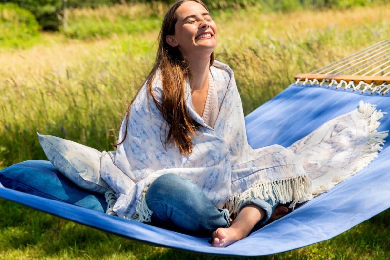 Brunette woman wrapped in an organic light blue blanket smiling into the sunlight while sitting corsslegged on a blue hammock