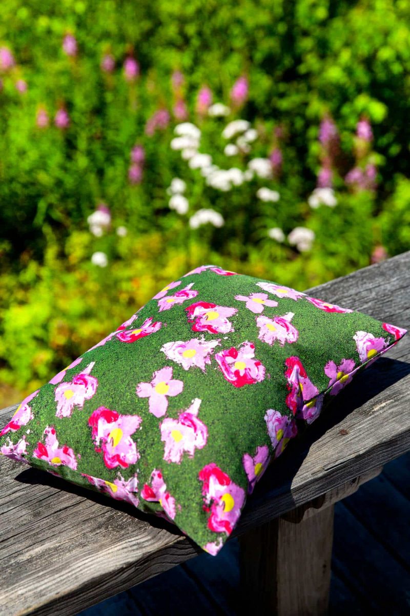Green organic pillow with pink flowers outdoors on a wooden railing with pink and white flowers in the background