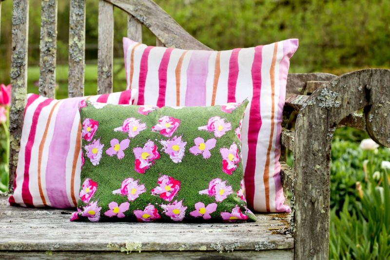 Two pink striped organic pillows and a green floral pillow on a rustic bench in a garden