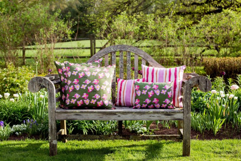 Two pink striped organic pillows and green floral pillows on a rustic bench in a garden