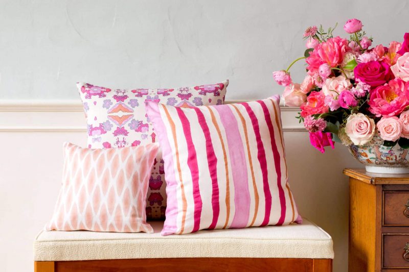 Three pink and orange organic pillows on a bench next to a pink floral arrangement