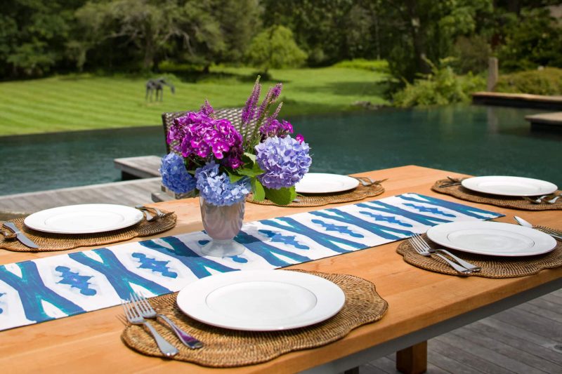 Poolside outdoor wooden dining table set with white plates wicker placemats purple flowers and organic table runner